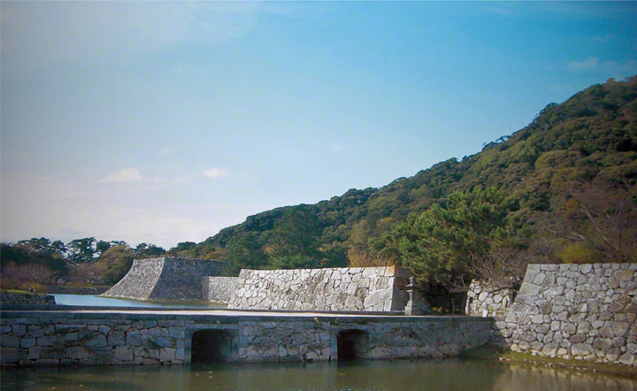 萩城跡。伝統的な防御のための石積み城壁と，指月山を背景に本丸橋の架かった内堀。本丸はその基礎の上に建っていました。